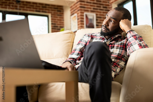 Thoughtful black man sits on couch, head resting on fist, gazing into distance with tired expression. African american freelancer reflecting on life, emotions and future decisions in quiet living room photo