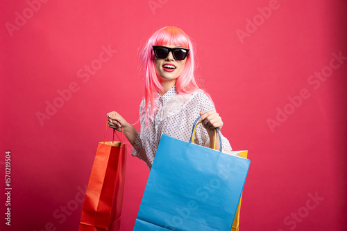 Happy young woman with pink hair and black sunglasses holding shopping bags. Stylish shopper in casual outfit, excited after seasonal sales and fashion discounts on isolated background.