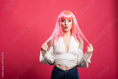 Confident woman in pink wig and off shoulder top poses with playful pout while holding strands of hair. Gorgeous model standing against isolated backdrop exuding quirky self assurance.