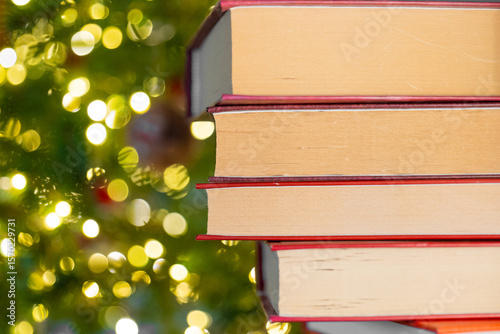 Books with red covers stack on Christmas tree background. Books on the shelf and fir branches with garlands. Literature with the atmosphere of winter and Christmas. 