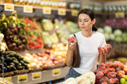 Woman near vegetable stand in store choose flat peach. Client view ripeness of fruit, take out fruits from showcase, collects grocery basket. Girl pull out ripest fruits from large pile of fruits.