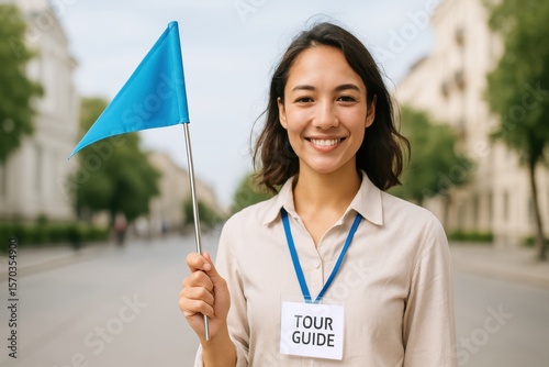 Cheerful young tour guide smiling and holding a blue flag while standing on a street with an urban background during a sunny day in a city environment