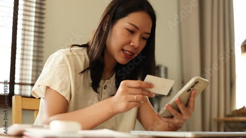 Young asian woman sitting in living room and use smartphone to making online transaction by credit card. Happy woman buying a new clothes and online payment for online shopping lifestyle concept