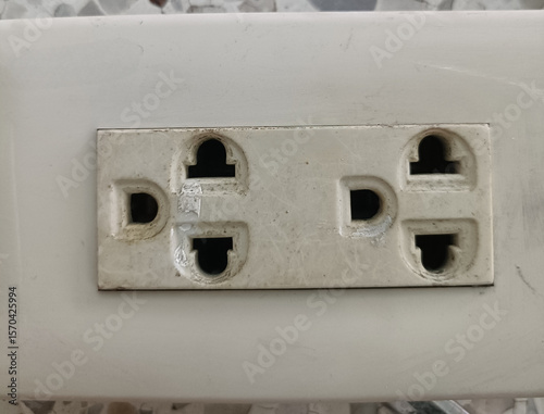 Close up of double electrical outlet with worn and dirty appearance, mounted on white surface, showing signs of age and use, with some dust and minor damage visible