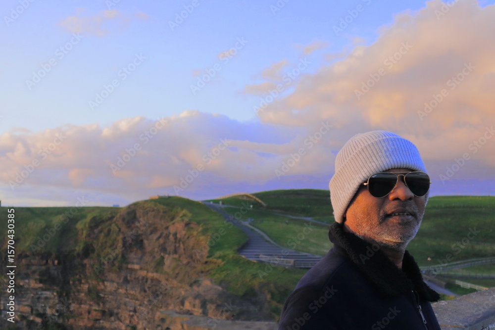 Naklejka premium Stylish Man at Cliffs of Moher on Bright Sunny Evening – Scenic View of Iconic Sea Cliffs and Atlantic Ocean in County Clare, Ireland with Clear Blue Sky and Dramatic Coastal Landscape