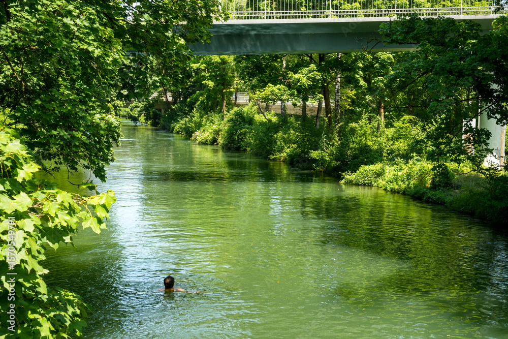 Obraz premium Floating man in the Eisbach (Isar), English Garden, Munich. Idyllic river landscape lined with lush greenery. A bridge spans the river. It is a tranquil and natural scene that invites you to linger