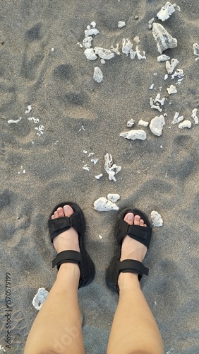 Feet in Black Sandals on Dark Sandy Beach with White Coral Fragments