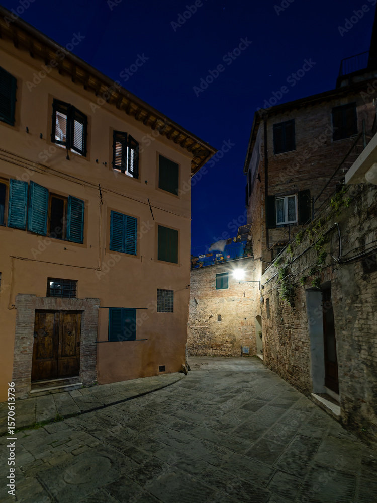 Fototapeta premium A small piazza in Siena, Italy, illuminated by warm streetlights under a dark blue night sky. The historic architecture, stone-paved square, and serene ambiance create a picturesque nighttime scene.