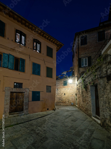 Fototapeta Naklejka Na Ścianę i Meble -  A small piazza in Siena, Italy, illuminated by warm streetlights under a dark blue night sky. The historic architecture, stone-paved square, and serene ambiance create a picturesque nighttime scene.