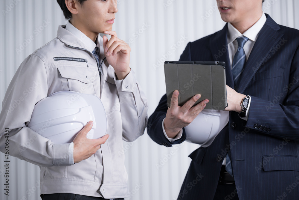 © kapinon - ゼネコンなどの現場で資料を確認する施工管理の作業服とスーツの男性　打ち合わせで確認する顔無しのバナー　考える