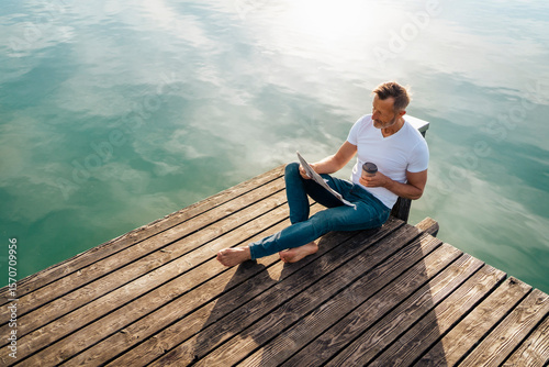 Man sitting with newspaper and disposable coffee cup on pier at lake