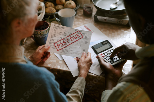 Senior Caucasian woman and senior Black man reviewing overdue utility bill with past due notice, holding paperwork and using calculator at kitchen table, focusing on financial stress