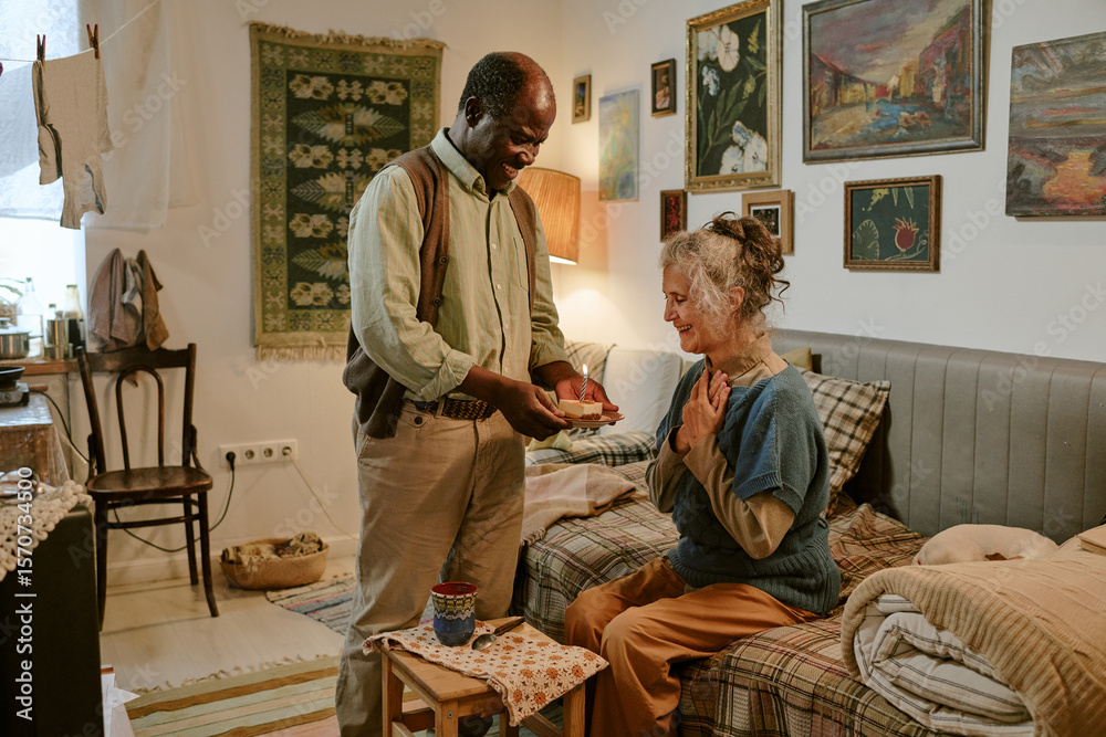 Fototapeta premium Senior Black man standing and giving small gift to senior Caucasian woman sitting on bed, both smiling warmly in modest room, elderly couple sharing heartfelt moment together