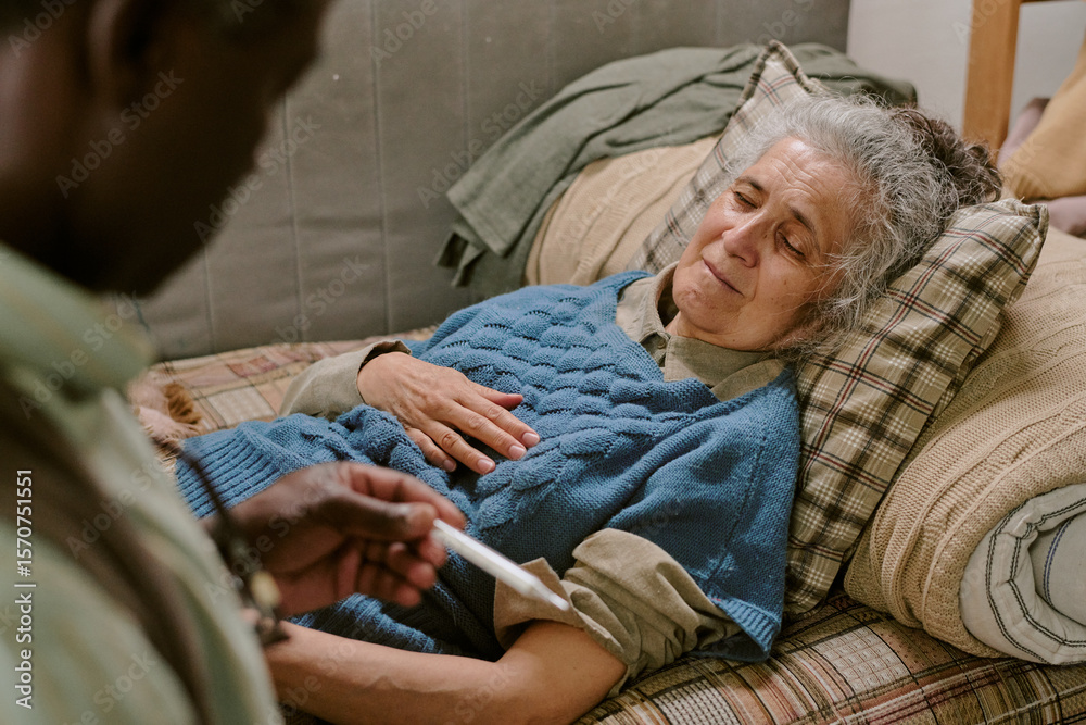Fototapeta premium Senior Caucasian woman lying on couch appearing unwell, senior Black man holding thermometer checking her temperature, both showing signs of concern and fatigue in modest home setting
