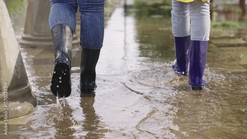 people walk along road in rubber boots through puddles, splashes fly from walking, wet asphalt road, flood city street, water spilling from banks, earth weather phenomenon, mud and puddles underfoot.