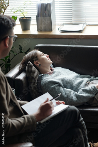 Woman lying on couch during therapy session with man taking notes, both seated in professional office setting with window blinds