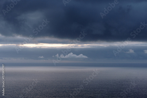 Dark storm clouds gather over a calm, gray ocean with a bright line of light on the horizon