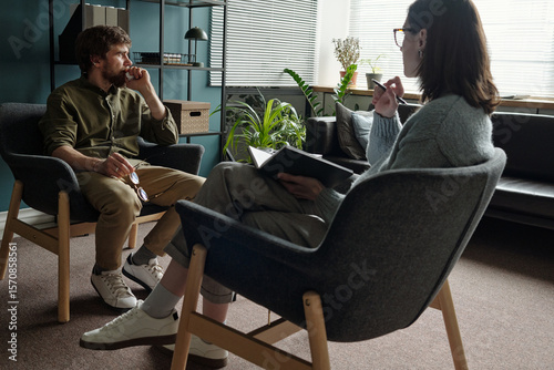 Man sitting in armchair talking to woman therapist holding notebook during therapy session in modern office setting