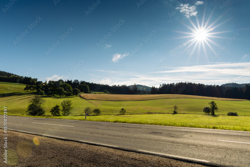 Fototapeta premium Foothills of the Austrian Alps with asphalt road on foreground and sun on blue morning sky