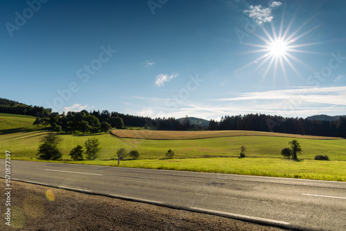Foothills of the Austrian Alps with asphalt road on foreground and sun on blue morning sky