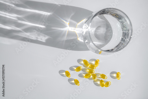 Yellow vitamin D or fish oil capsules are scattered near a glass of water, casting intriguing shadows on a white surface, creating an aesthetically pleasing composition