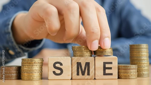 Coins Stacked with Wooden Cubes Spelling Sme with Hand and Blue Shirt on Bright Surface