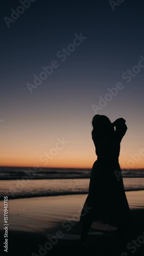 Silhouette of a woman dancing gracefully on the beach as the sun sets. Her movements are fluid and elegant, creating a captivating visual against the backdrop of the ocean and the colorful sky.