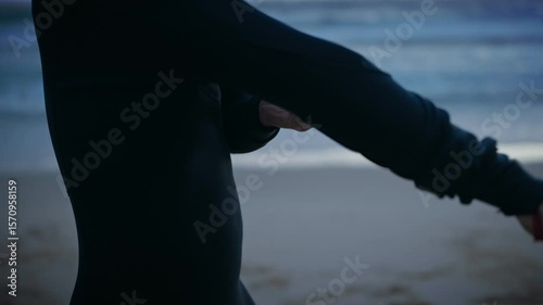 A surfer puts on their wetsuit, preparing to enter the ocean and surf. They are on the beach at dawn or dusk, and the waves are small.