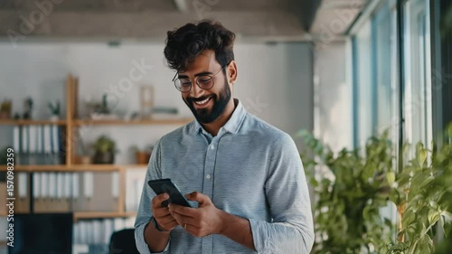 A young Hispanic man uses a phone application while perusing web sites in his office. A happy worker makes a call and dials a message.