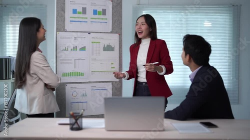 Three people are standing in front of a board with graphs on it. One of the women is wearing a red jacket