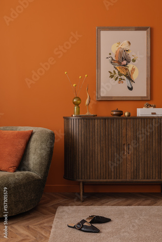 Warm and eclectic interior with a retro-style olive armchair, wooden sideboard, mock up art print on orange wall. Soft lighting enhances the cozy mood.
