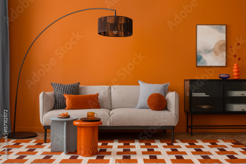 Modern living room with orange wall, curved floor lamp and cozy grey sofa with colorful pillows. Mock up poster frame.