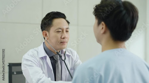 Asian male doctor using stethoscope to examine patient’s chest during physical checkup in hospital setting, healthcare professional diagnosing breathing or heart condition in clinical.
