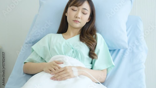 Young Asian woman resting in hospital bed with IV catheter in hand, wearing hospital gown, peaceful expression, receiving medical treatment or recovering from illness in a clean clinical.