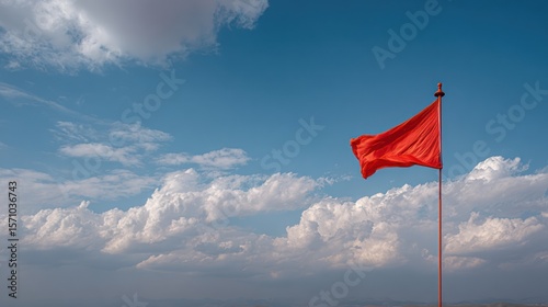 Bright red flag flying atop a tall pole under a blue sky filled with puffy clouds