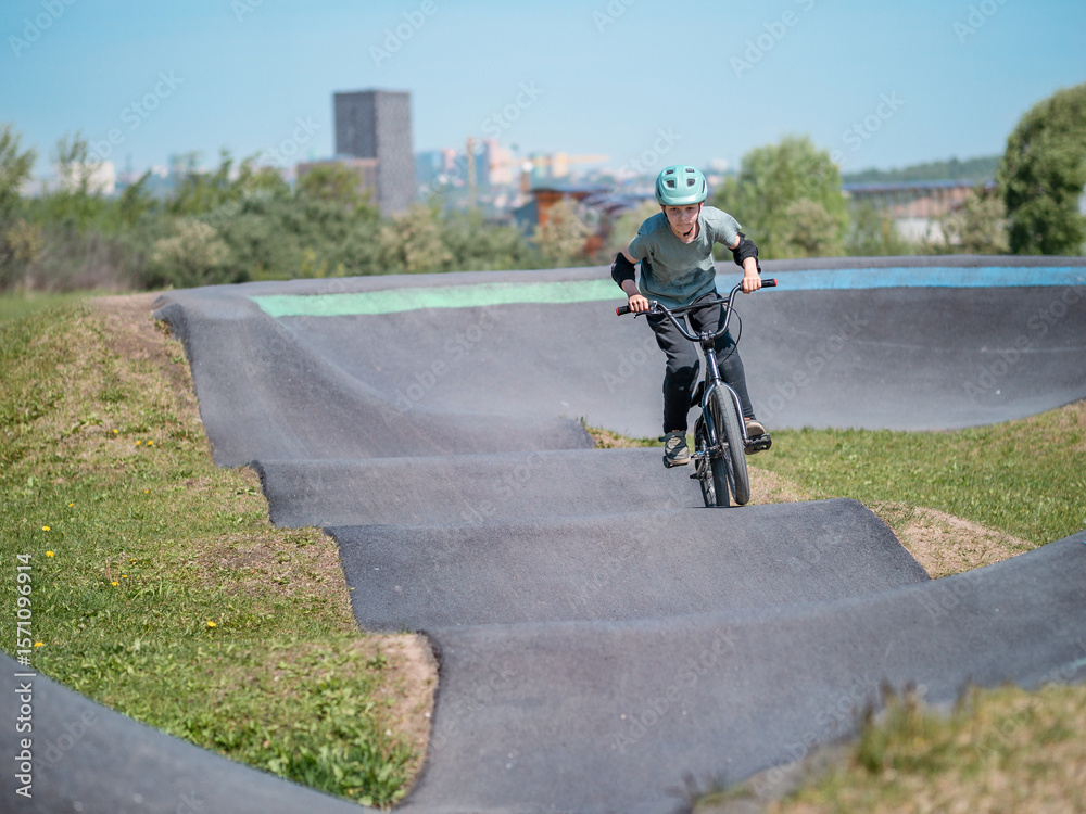 Fototapeta premium Child on bmx bike on pump track. Young male cyclist in helmet and protection rides bmx bike on asphalted bicycle race track. Boy BMX rider having fun and training on racing speed track. Extreme sport