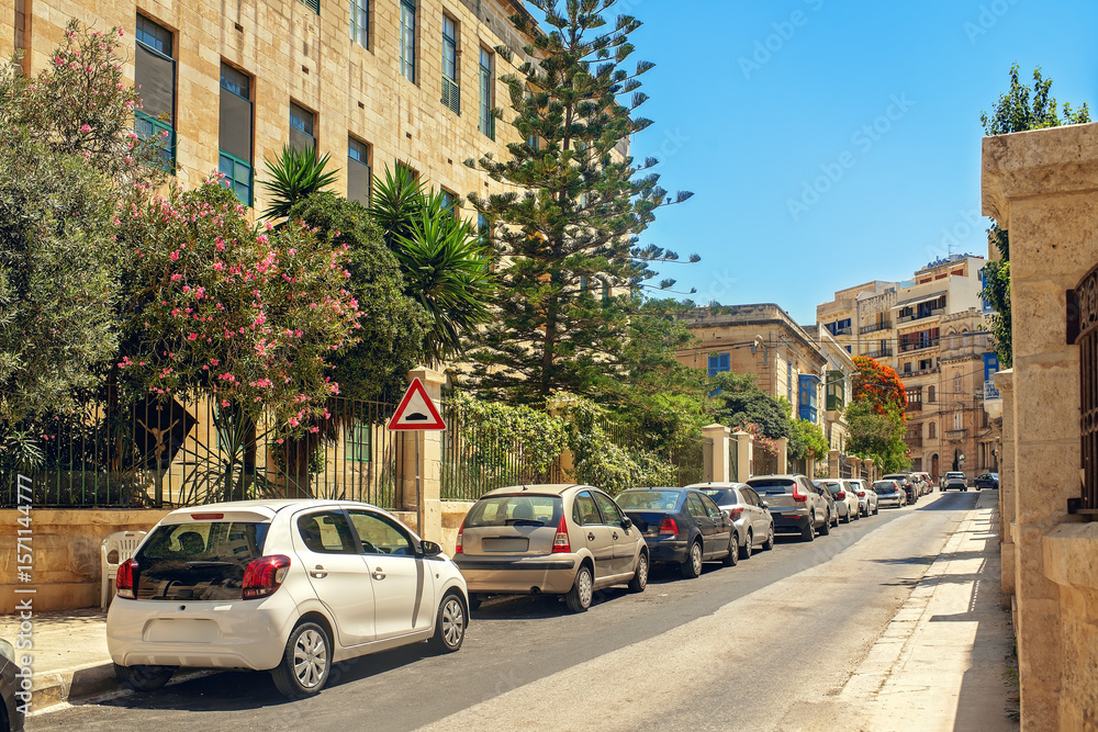 Obraz premium Quiet European street with parked cars along a metal fence, tropical plants, old buildings, and a road sign. Concept of summer, travel, and everyday urban life.