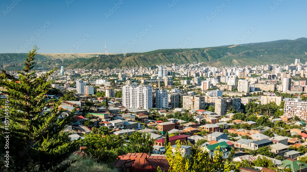 Fototapeta premium Panoramic view of Tbilisi cityscape with green hills and clear blue sky, capturing urban life against natural mountain backdrop in Georgia
