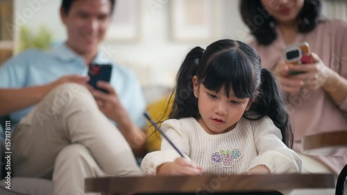 Young Asian girl drawing while her parents using smartphones not engaging with their daughter at home.child drawing while her parents sit on sofa.Concept of family disconnection,Digital Distraction