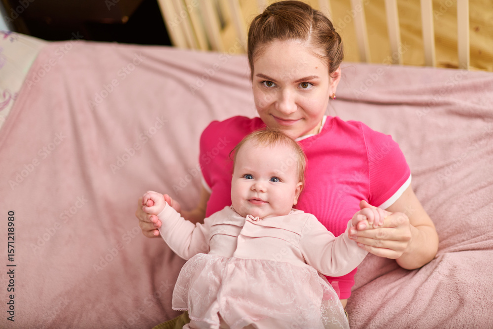 Young adult female with brown hair holding adorable baby girl in pink outfit. European ethnicity, both smiling indoors on soft pink blanket with wooden crib nearby Young adult female with brown hair holding adorable baby girl in pink outfit. European ethnicity, both smiling indoors on soft pink blanket with wooden crib nearby
