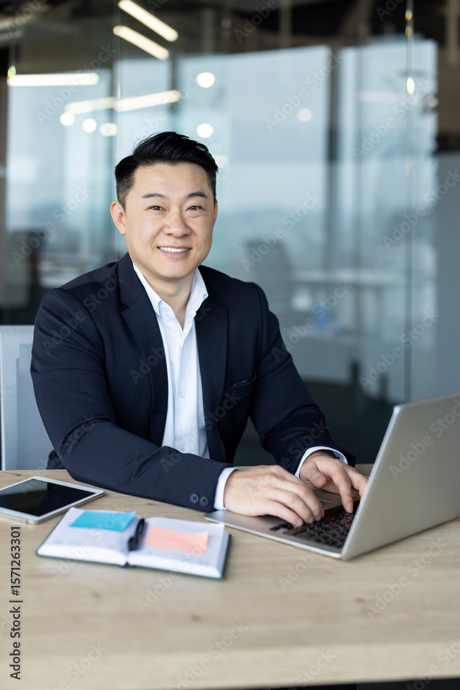Vertical photo of a smiling and successful young Asian businessman sitting at a desk in the office working on a laptop Vertical photo of a smiling and successful young Asian businessman sitting at a desk in the office working on a laptop