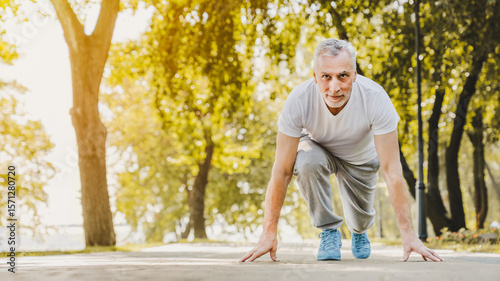 Athletic fitness senior man starting running race in sunny park. Active elderly male jogging in summer forest during morning workout outdoors. Active retirement, sport healthy lifestyle concept