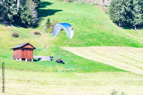 Dynamic shot of paragliding in the Italian Alps. A vibrant paraglider hovers over a green meadow next to a rustic wooden cabin. Adventure sport in nature.