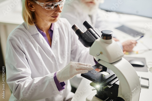 Caucasian middle aged woman examining specimen under microscope in scientific laboratory, wearing protective eyewear and gloves, older Caucasian man working in background
