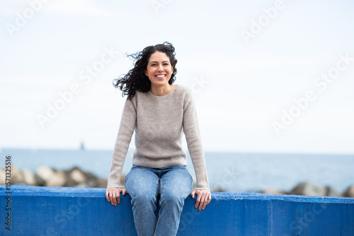 Vászonkép Portrait smiling woman sitting on blue concrete ledge by the sea