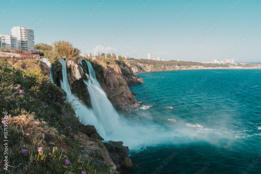 Obraz premium Scenic view of Duden Waterfalls in Antalya, Turkey, cascading from rocky cliffs into the Mediterranean Sea, with blooming purple flowers in the foreground and a turquoise sky.