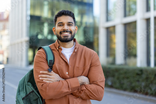 A smiling Indian student with a backpack stands confidently outside a modern building, with arms crossed