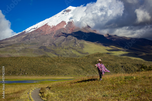 Woman in traditional attire stands admiring the breathtaking view of cotopaxi volcano. The scene captures the vibrant contrast between lush greenery and the snow-capped peak