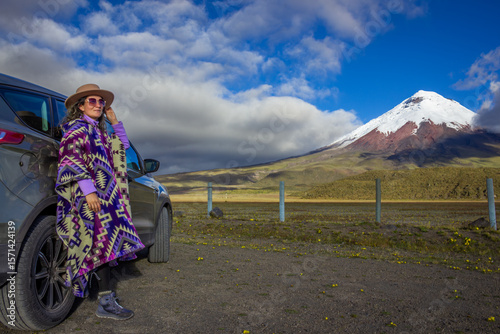 Stylish tourist wearing a poncho and hat is posing next to her suv car during a road trip in the scenic cotopaxi national park, ecuador, with the majestic cotopaxi volcano in the background