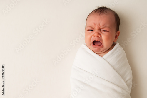A tightly swaddled newborn baby crying loudly, lying on a soft surface with arms tucked inside the wrap. The baby's face is red with distress, expressing discomfort and need for attention. The image o